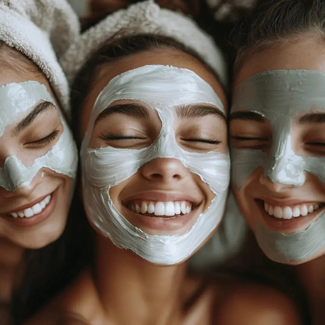 Three smiling women with closed eyes wearing white and grey clay face masks, two with towels on their hair